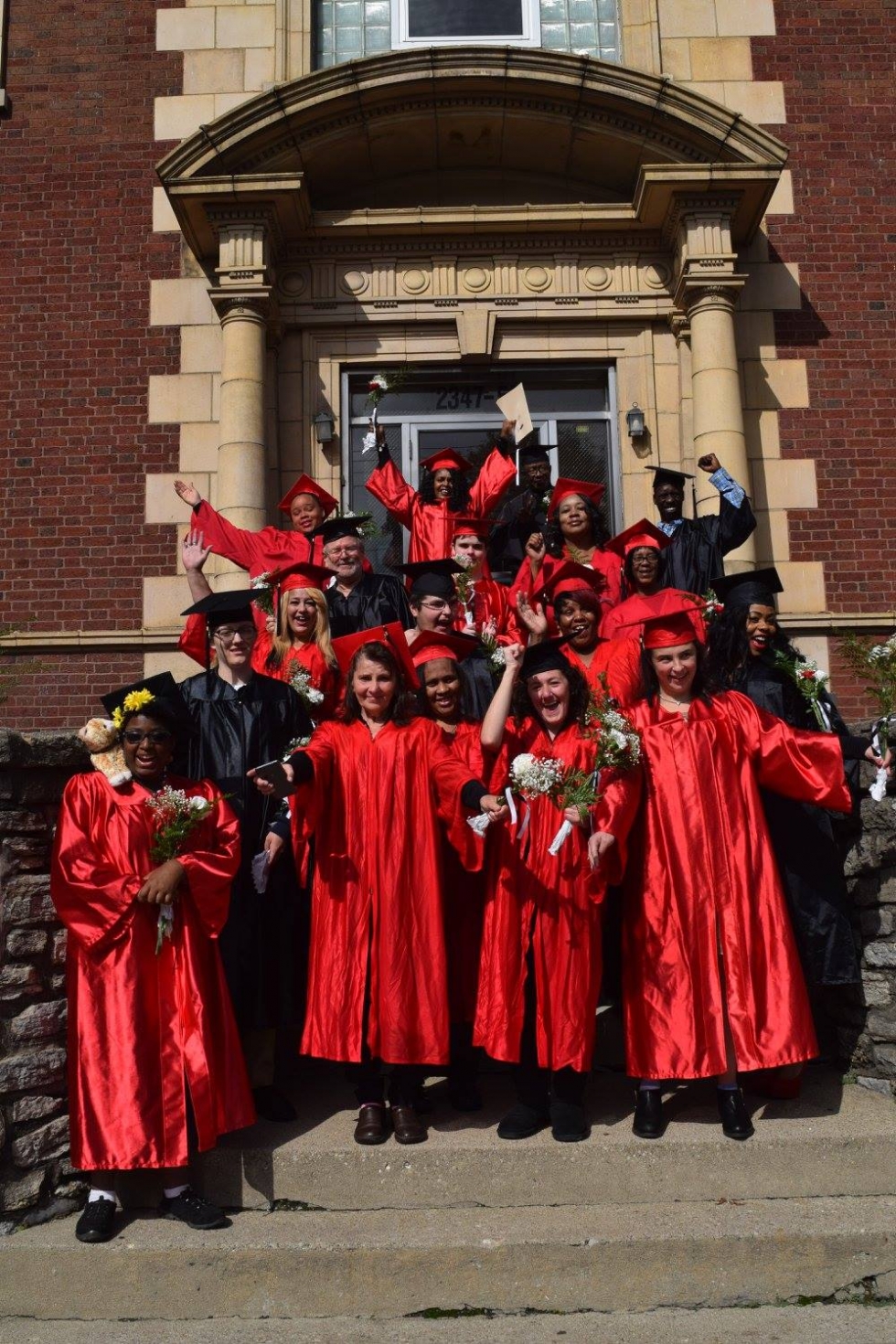 A group of people in graduation robes stand on the front steps of the IKRON building. Graduates are cheering, have arms extended, and look happy and celebratory.